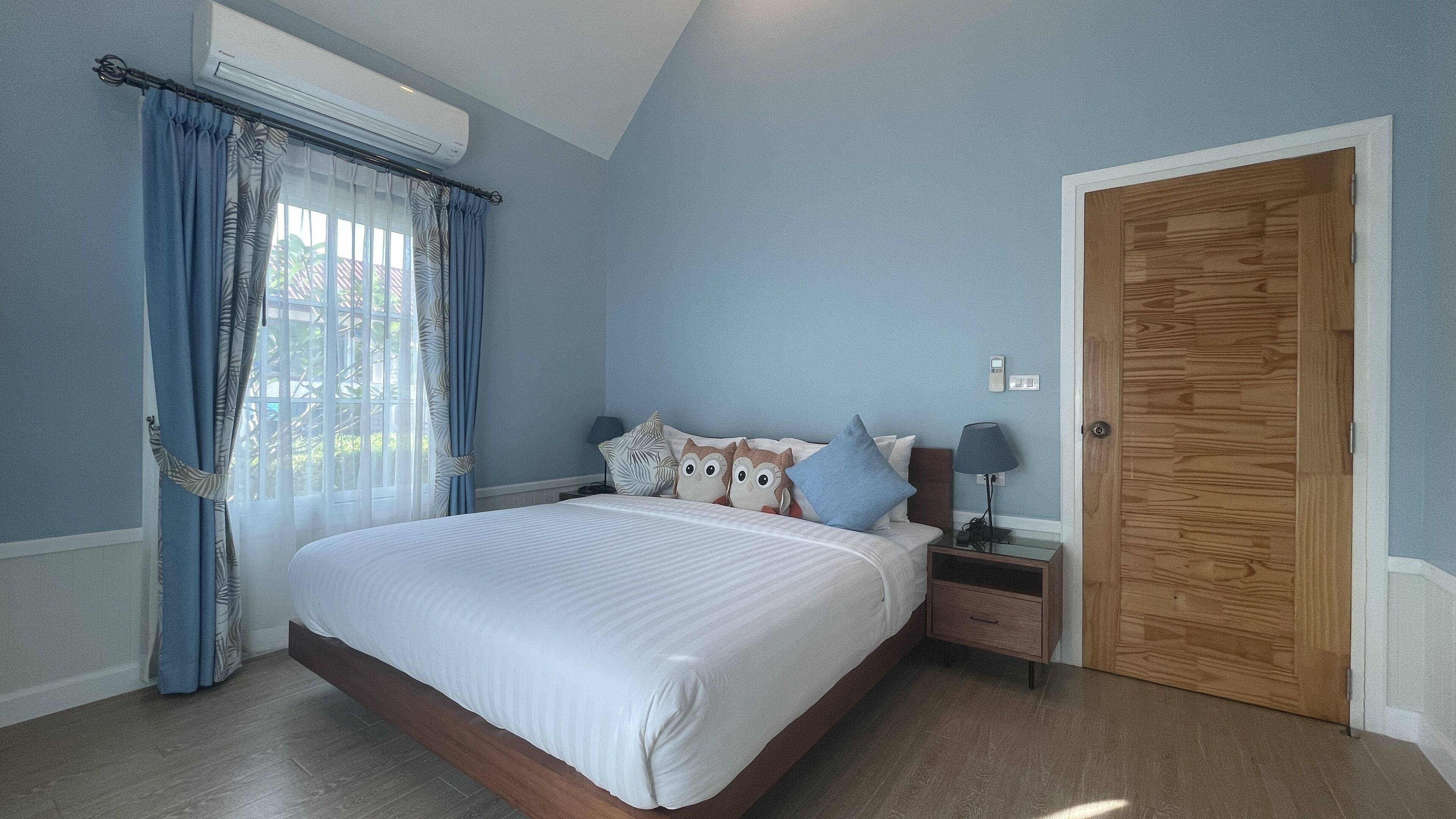 Un dormitorio con cama, mesitas de noche, lámpara, puerta de madera y ventana con cortinas.