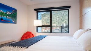 A hotel room with a large bed, a window with a mountain view, a painting on the wall, and a red chair. - Hygge Haus By Revelstoke Vacations (Revelstoke)