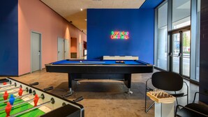 A pool table with a neon sign, a foosball table, and a chess table in a room with large windows and a blue accent wall. - CX Venice Mestre (Mestre)