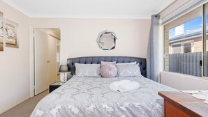 A neatly arranged bedroom with a large bed, a bedside table, a mirror, and a view of the outside. - Our Beach House- Busselton (West Busselton)