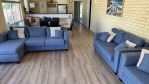 A living room with a blue sectional sofa, a kitchen area with a brick wall, and a painting of a coastal scene. - Our Beach House- Busselton (West Busselton)