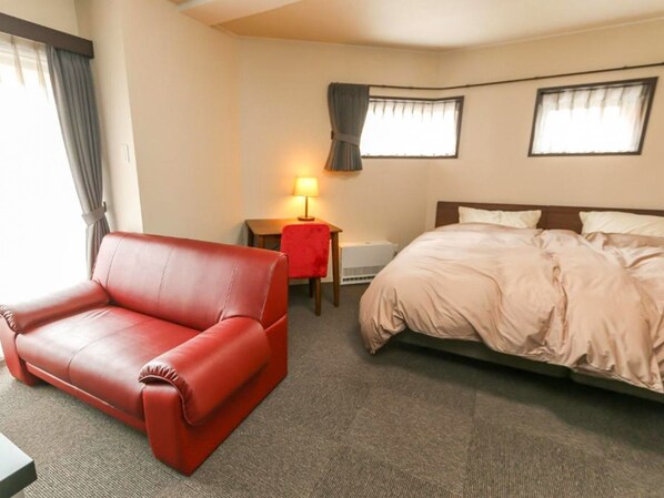 Habitació d'hotel amb un sofà de cuir vermell, un llit amb edredó beix, un escriptori amb una làmpada i dues finestres amb cortines.