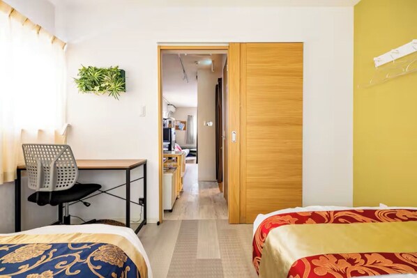 Ein Hotelzimmer mit zwei Betten, einem Schreibtisch mit Stuhl, einer kleinen Küchenzeile und einer Holztür zu einem weiteren Zimmer.