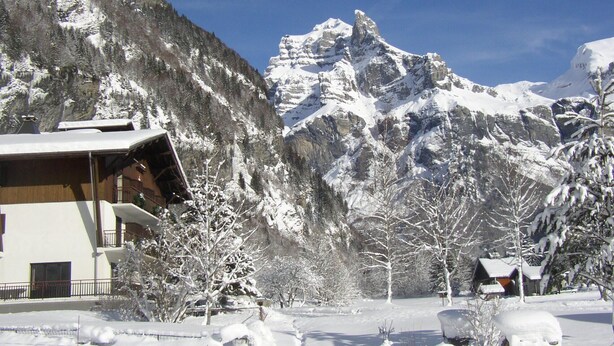Un albergue de montaña con fachada de madera, árboles cubiertos de nieve y un imponente pico montañoso al fondo.