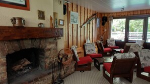 Una habitación acogedora con chimenea de piedra, paredes de madera y suelo de baldosas. Hay varias sillas y una mesa con un cojín rojo.