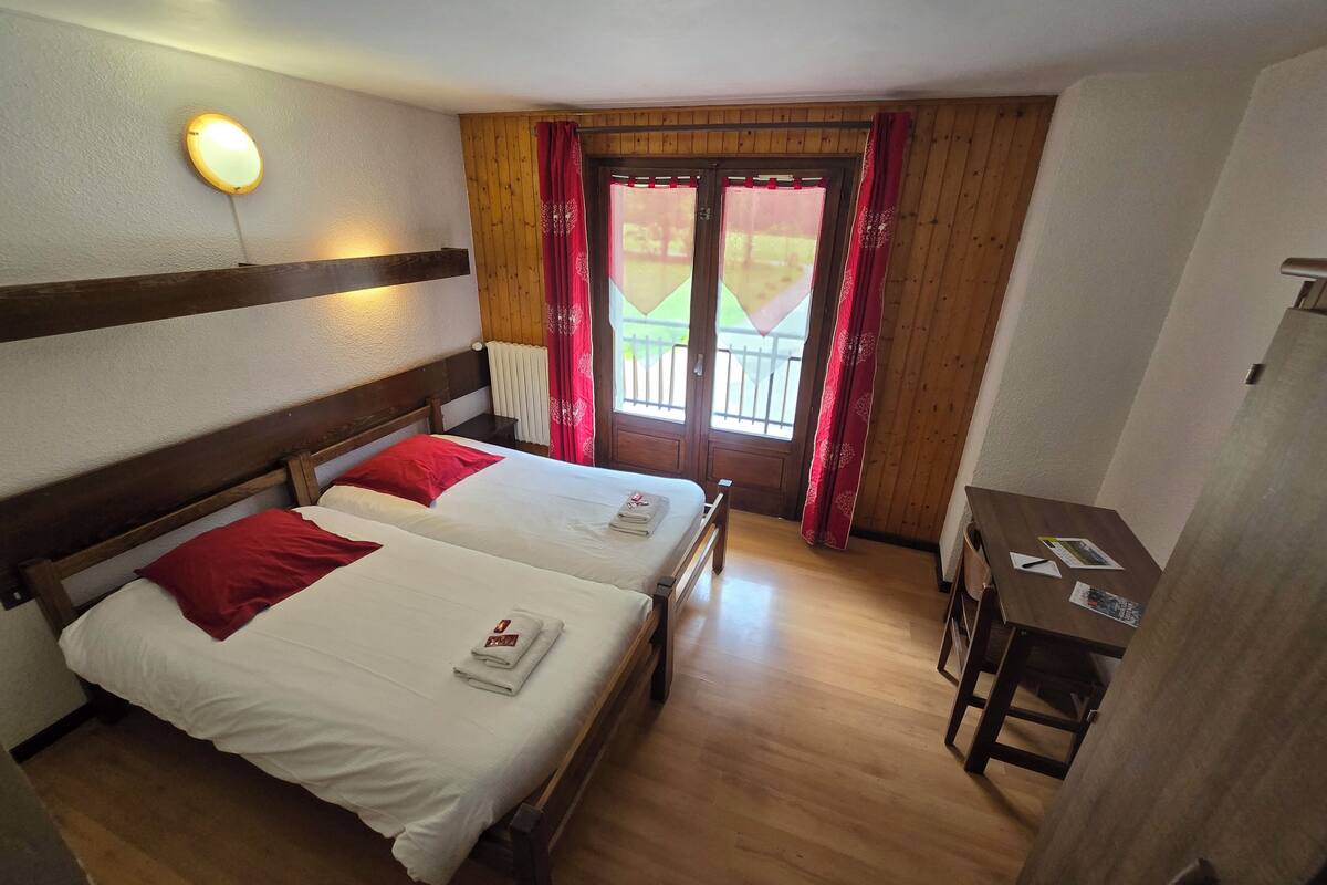 Une chambre d’hôtel avec deux lits en bois, un bureau en bois et une fenêtre ornée de rideaux rouges.