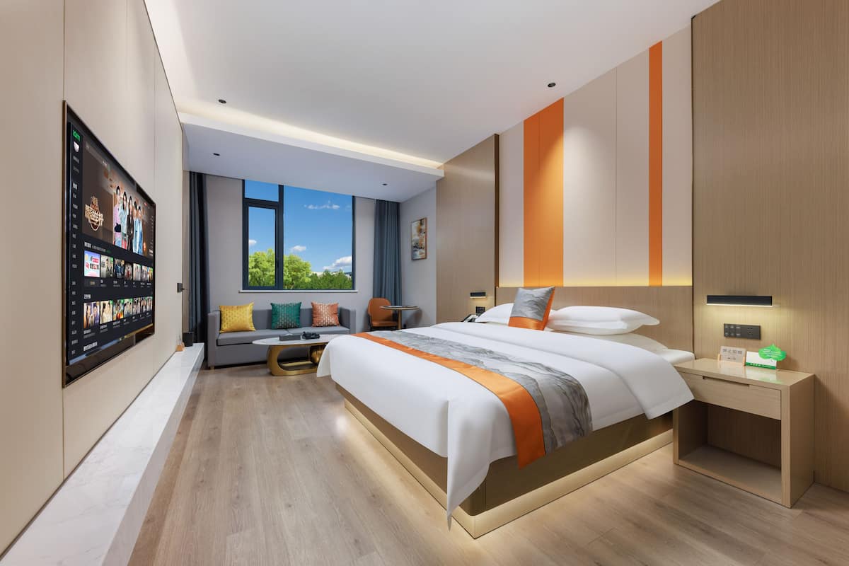 Une chambre d’hôtel moderne dotée d’un grand lit, d’un bureau, d’un coin salon avec un canapé et des fauteuils, d’un téléviseur fixé au mur et d’une fenêtre donnant sur un paysage verdoyant.
