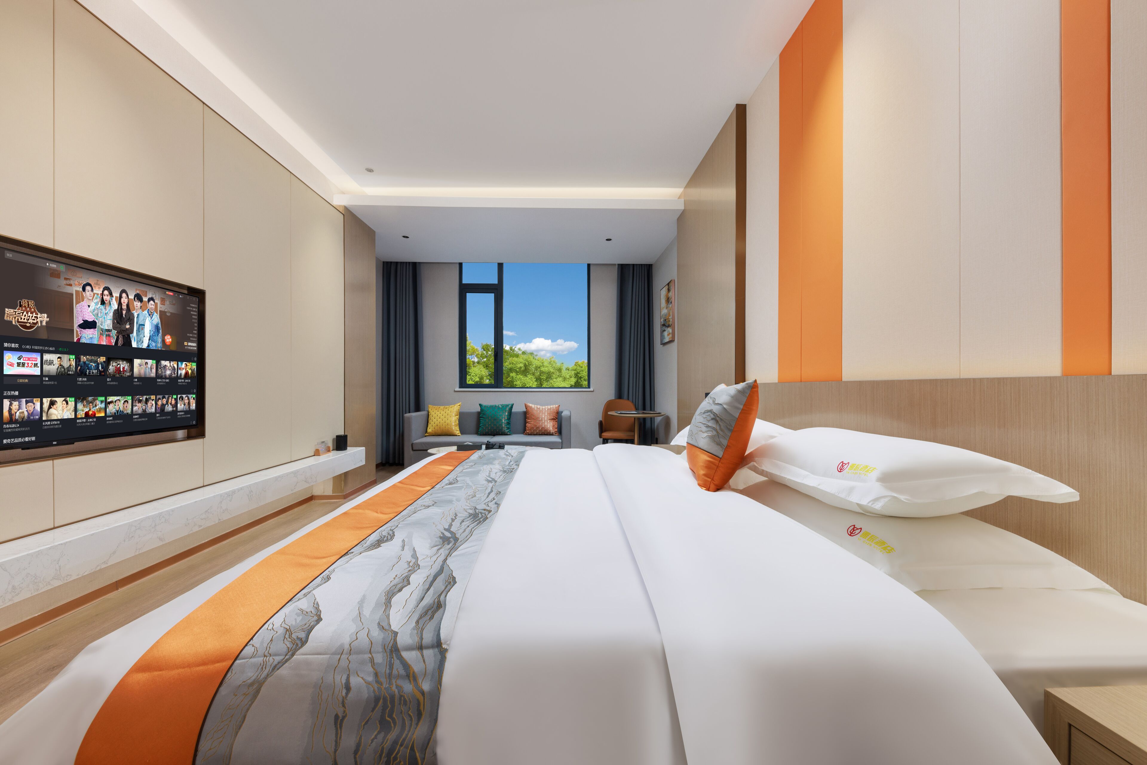A modern hotel room with a large bed, a flat-screen TV mounted on the wall, a sofa, and a window with a view of greenery.