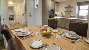 A dining area with a wooden table set for four, surrounded by brown leather chairs. A fruit bowl with apples and pears is on the table, and there is a kitchen in the background with modern appliances. - Thorneydyke Cottage (Stirling)