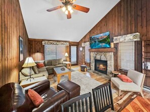 A cozy living room with a fireplace, wooden walls, a ceiling fan, and a television.