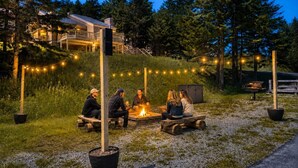 A group of people sitting around a fire pit in a natural setting with trees and a building in the background.
