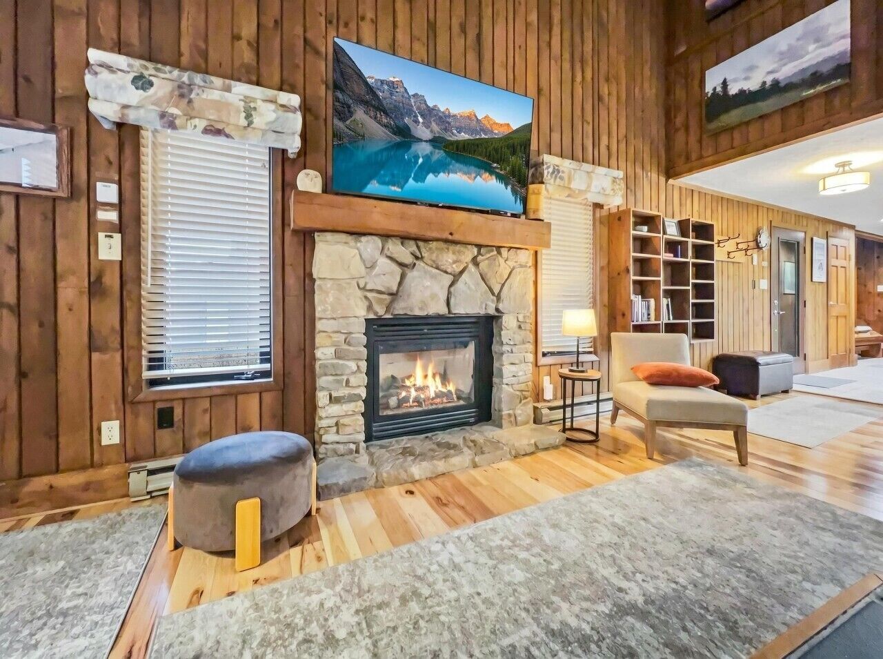 A cozy living room with a stone fireplace, a large flat-screen TV mounted above it, and a comfortable seating area with a sofa and ottoman.