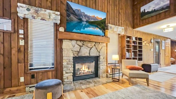 A cozy living room with a stone fireplace, a large flat-screen TV mounted above it, and a comfortable seating area with a sofa and ottoman.