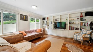 Ein Wohnzimmer mit einer Couch, einem Hocker und einem Fernseher auf einem Schrank. Es gibt Bücherregale mit verschiedenen Gegenständen und ein Fenster mit Jalousien.