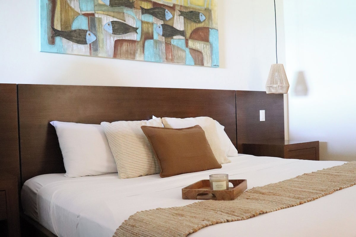 Cama bem arrumada com cabeceira de madeira, travesseiro marrom e travesseiro branco. Há uma pequena bandeja de madeira com uma vela sobre a cama.