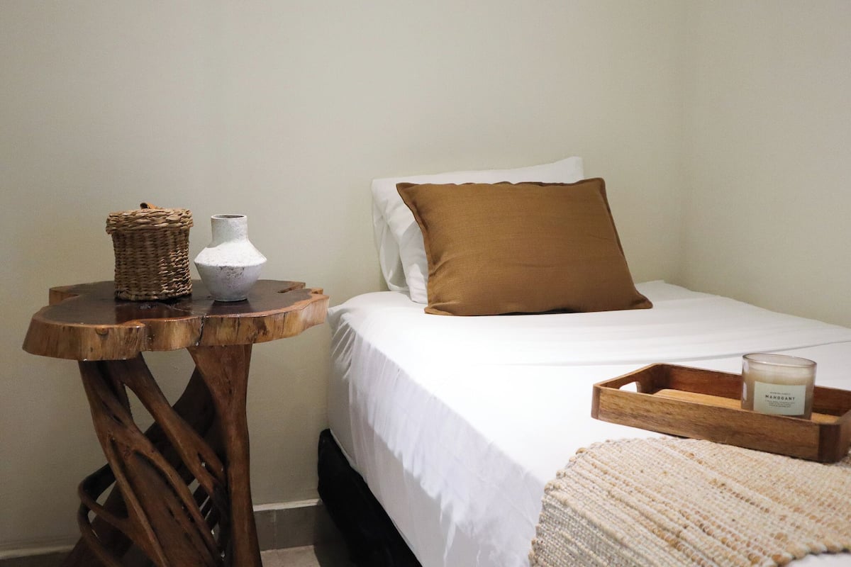 Quarto com mesa de cabeceira de madeira, vaso branco, travesseiro marrom, colcha branca e bandeja de madeira com uma vela.