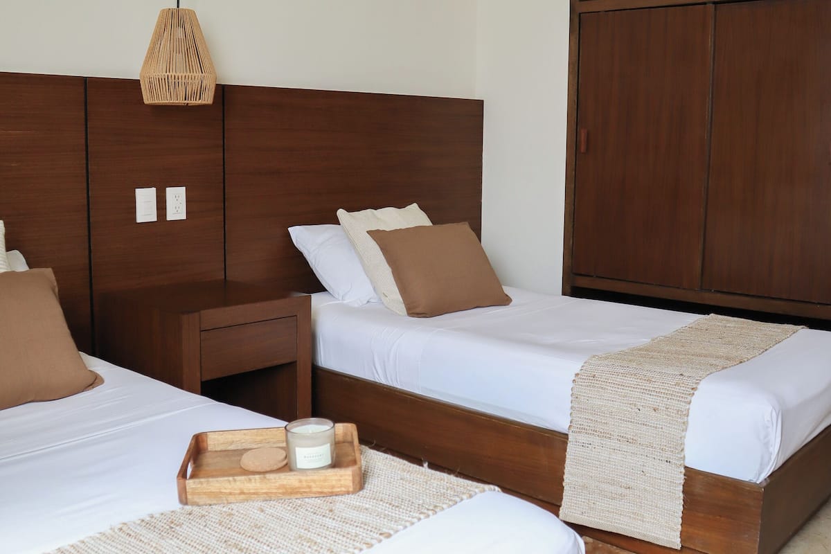 Quarto de hotel com duas camas, cabeceiras de madeira, mesa de cabeceira, luminária de tecido e um guarda-roupa espaçoso.