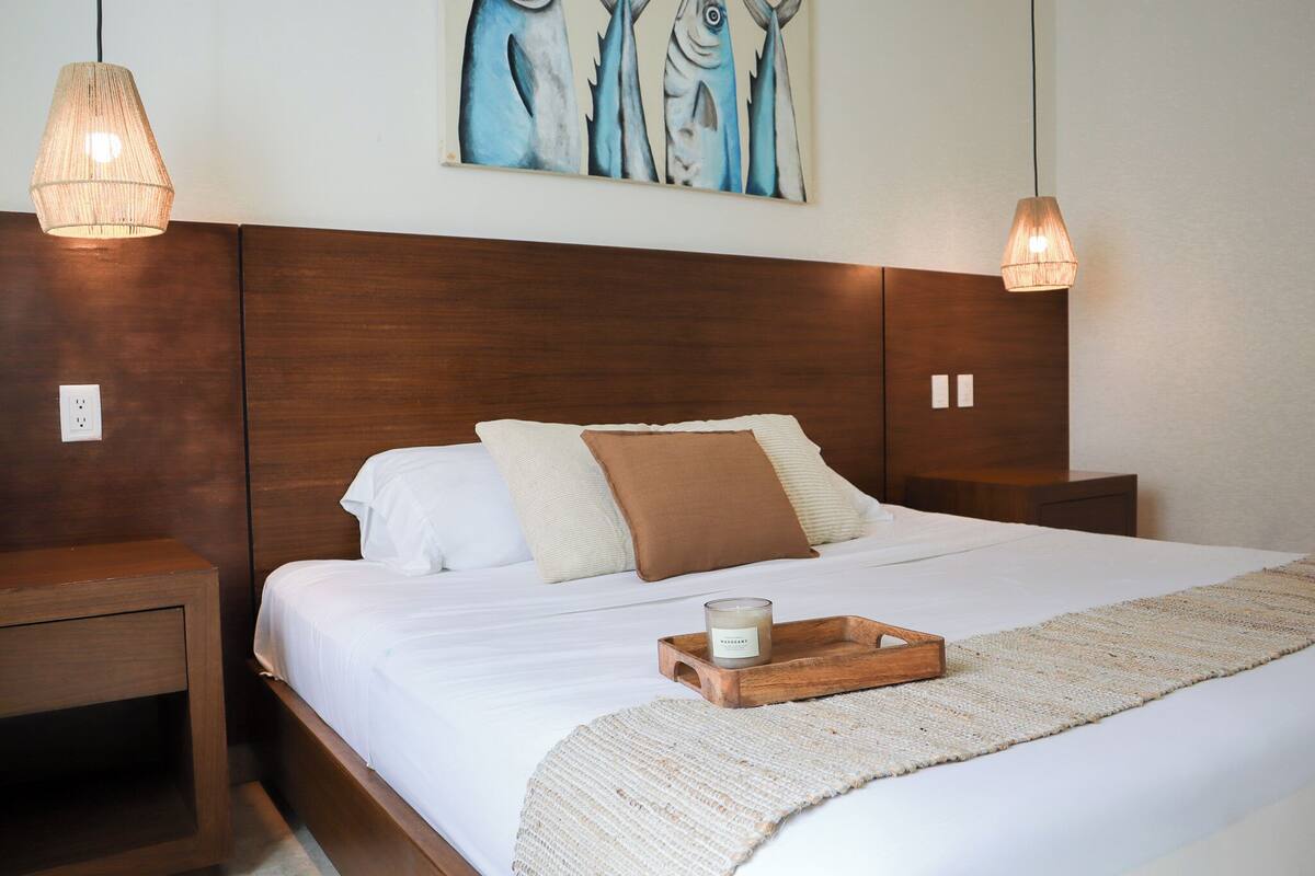 Cama bem arrumada com lençóis brancos, um travesseiro marrom e uma bandeja de madeira com uma vela. Há um quadro com peixes na parede acima da cama.