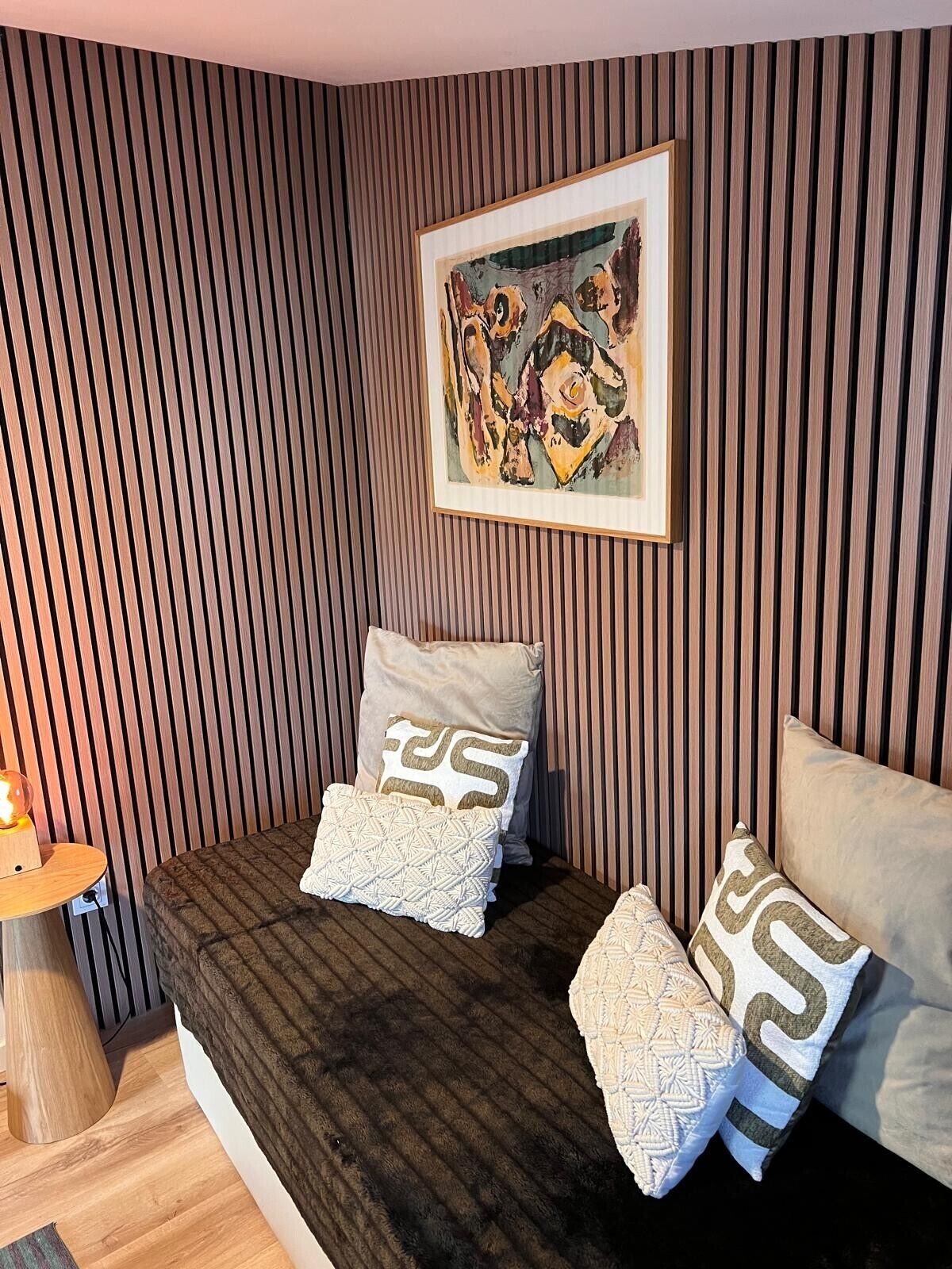 Een kamer met een lambrisering, een ingelijst schilderij, een bed met bedkussens met een patroon en een nachtkastje met een lamp.