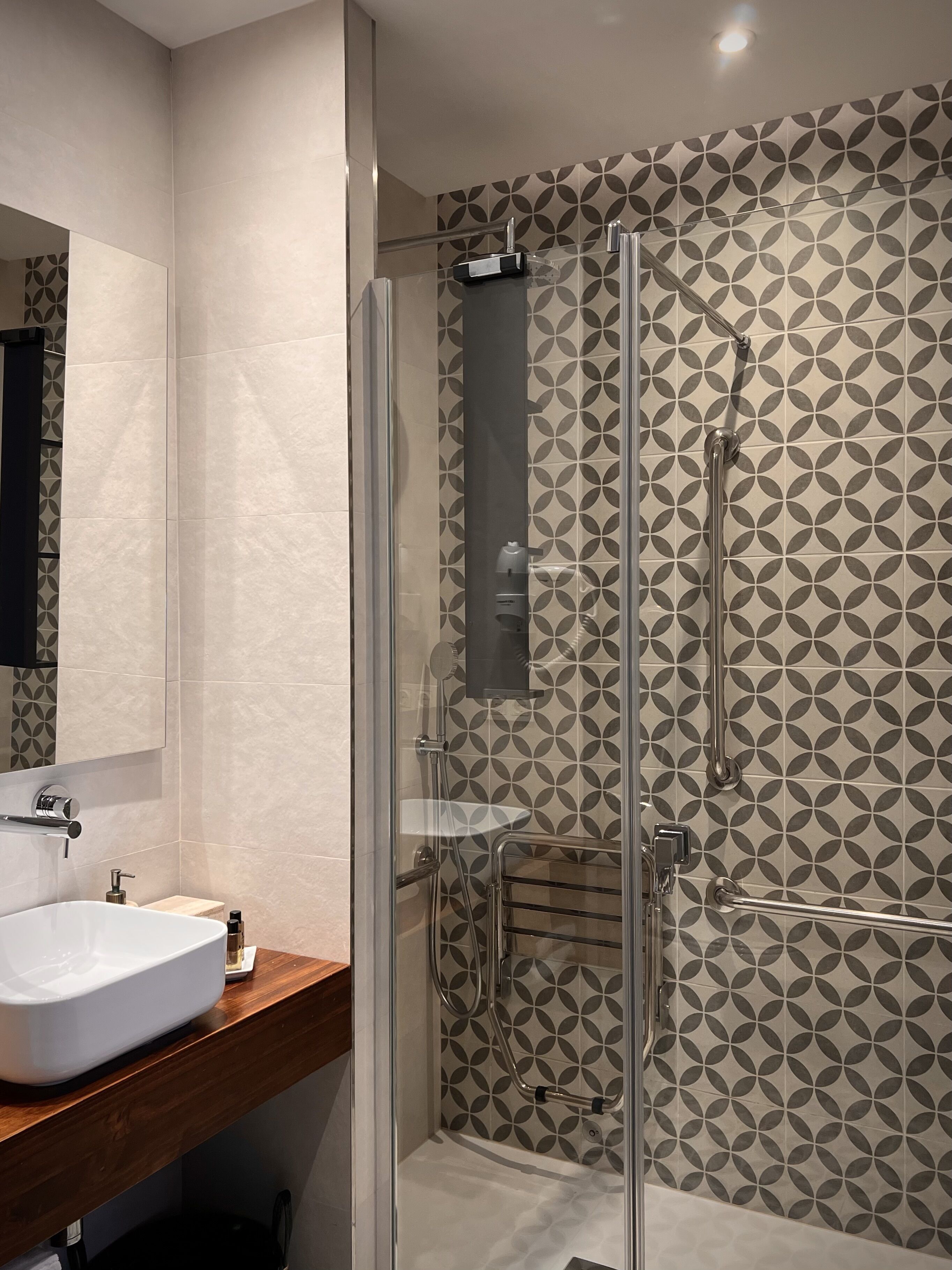 Casa de banho moderna com cabine de duche em vidro, toucador de madeira com lavatório branco e azulejos estampados nas paredes.