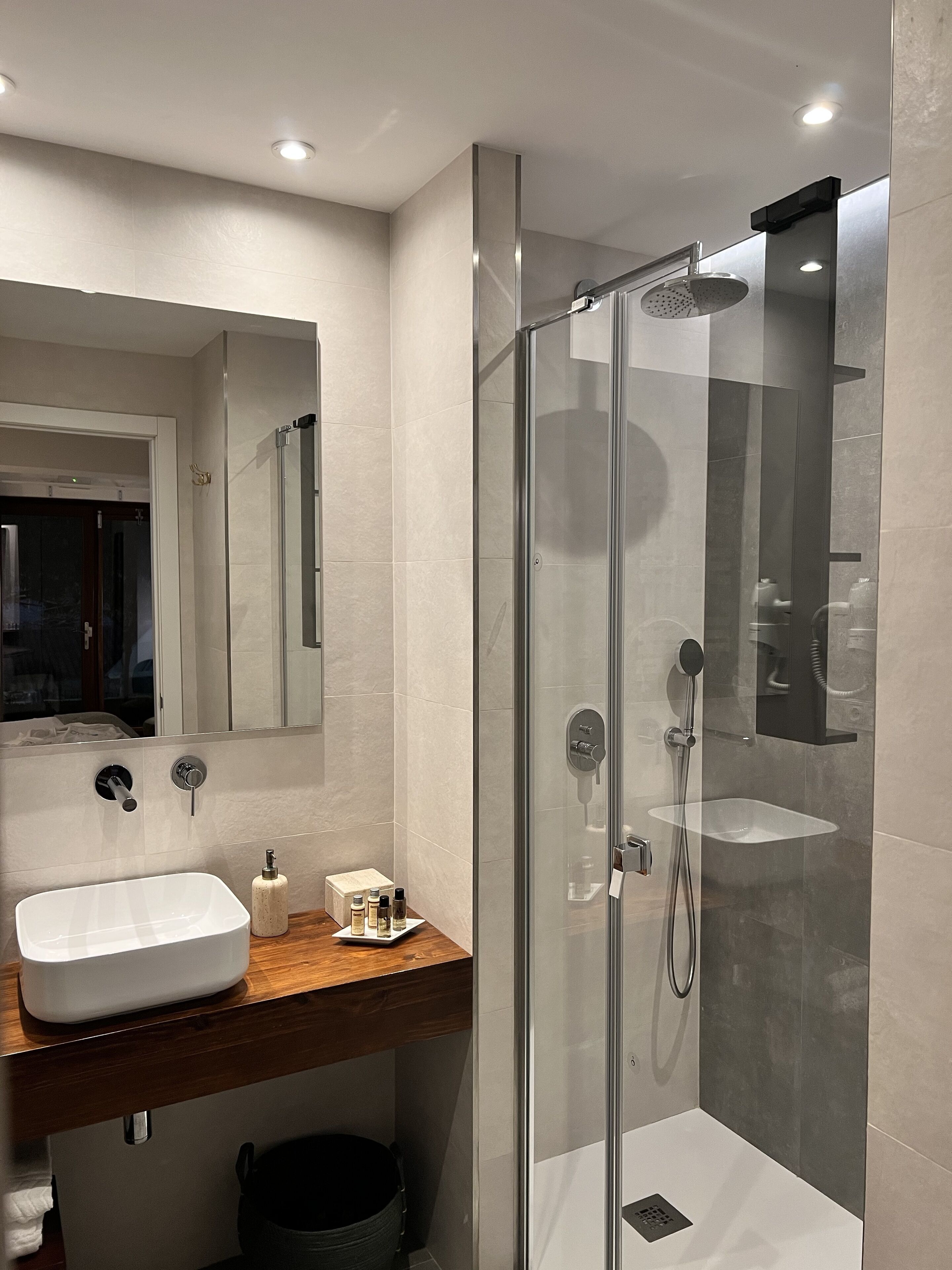 Un baño moderno con ducha envuelta en vidrio, un lavamanos blanco y un mueble de tocador de madera.