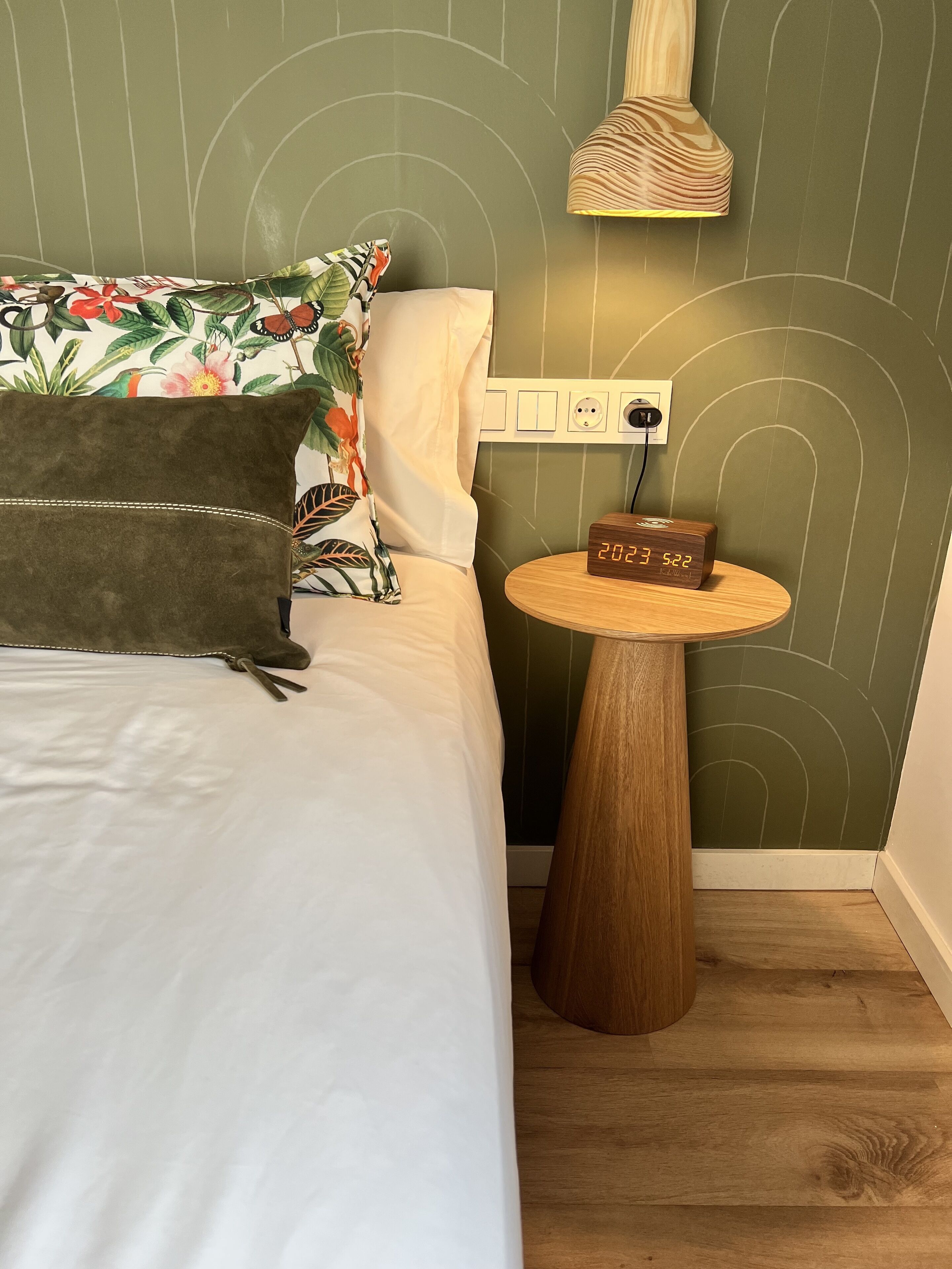 Una cama bien hecha con almohadones florales, una mesita de madera con un reloj y una lámpara de pared.
