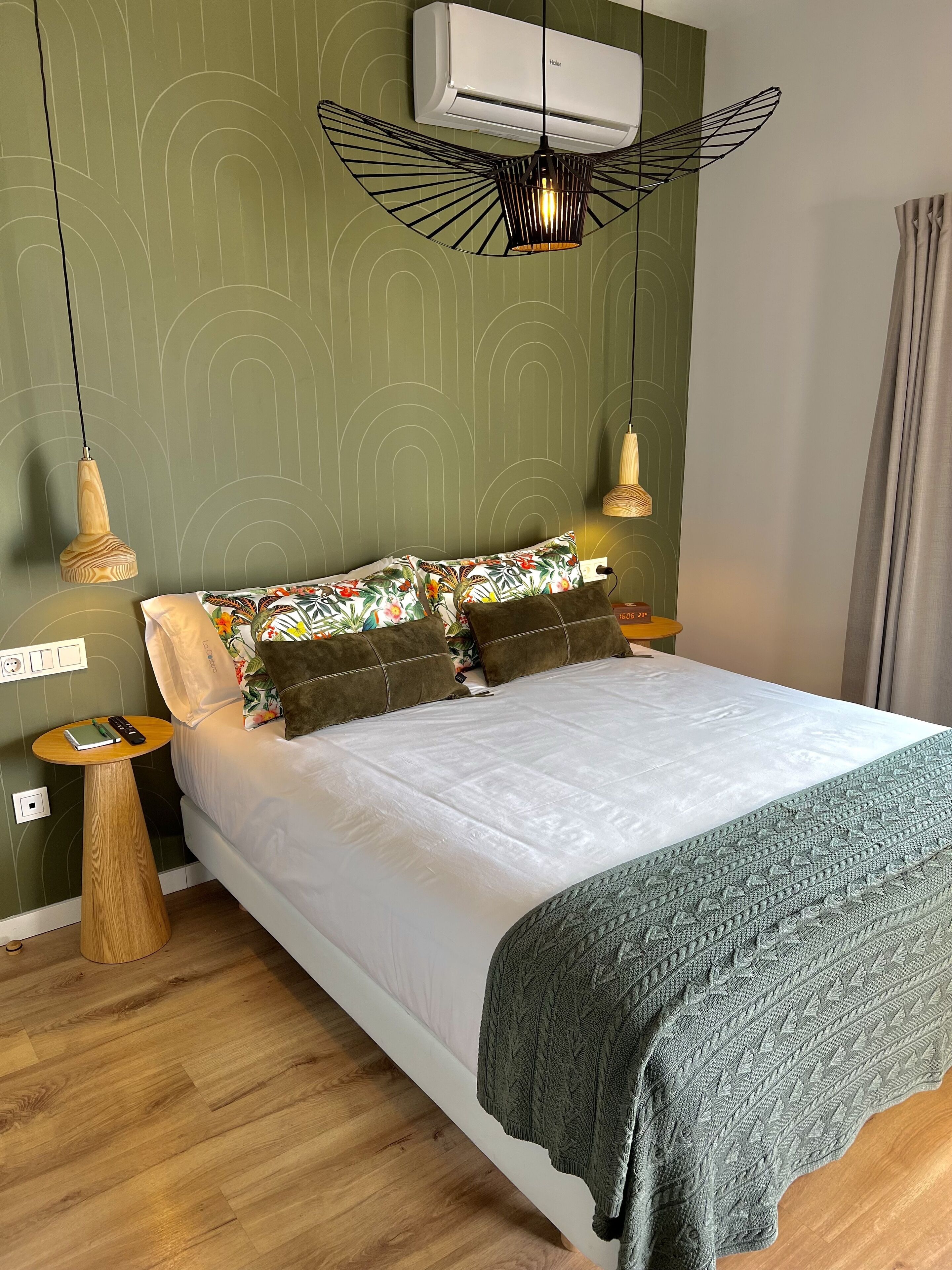 Una cama bien hecha con edredón blanco y manta verde de punto, una mesita de madera con un cuaderno y un bolígrafo, y un aire acondicionado instalado en la pared.
