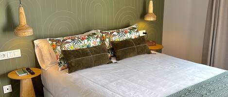 Una cama bien hecha con edredón blanco y manta verde de punto, una mesita de madera con un cuaderno y un bolígrafo, y un aire acondicionado instalado en la pared.