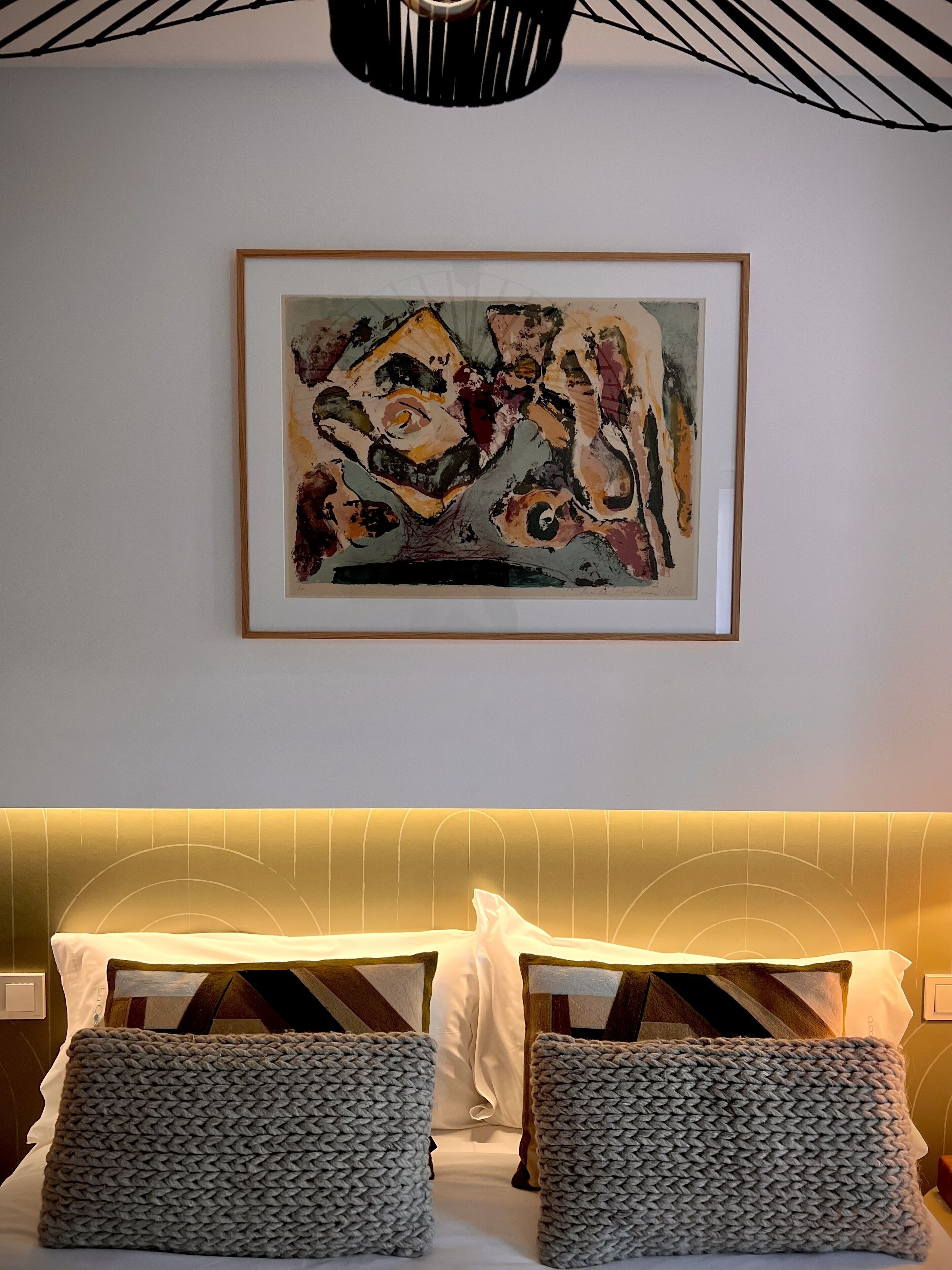 Een hotelkamer met een bed, twee grijze gebreide plaids, een gekaderd abstract schilderij aan de muur en een moderne plafondlamp.