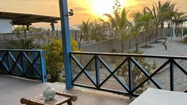 A covered outdoor seating area with a view of the sunset, a wooden chair, a small table with a decorative item, and a white cushioned bench. - Casa de Playa Cahuve Frente al mar (Cardalito)