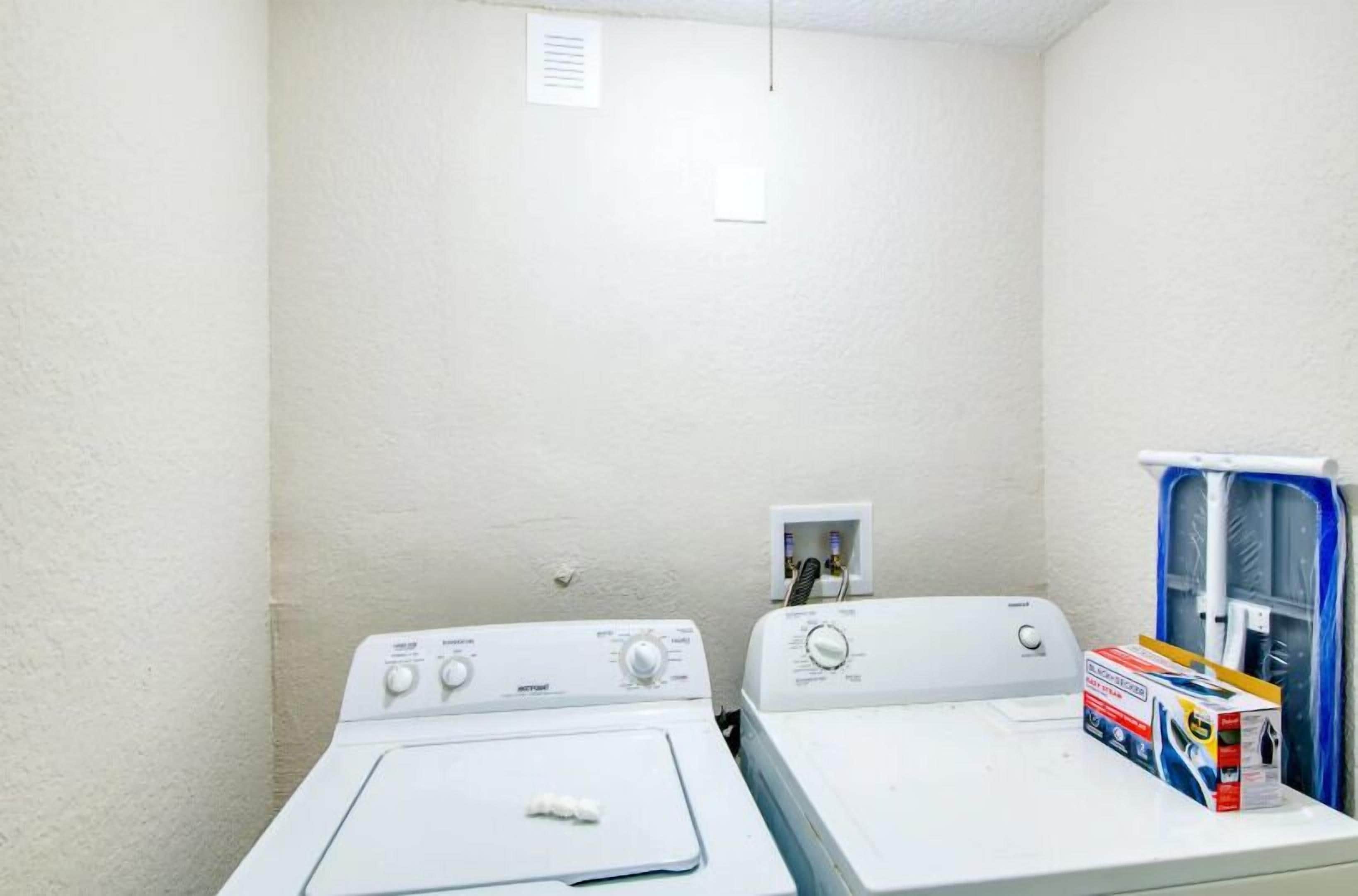 A laundry room with two white washing machines, a power outlet, and a window with a curtain.
