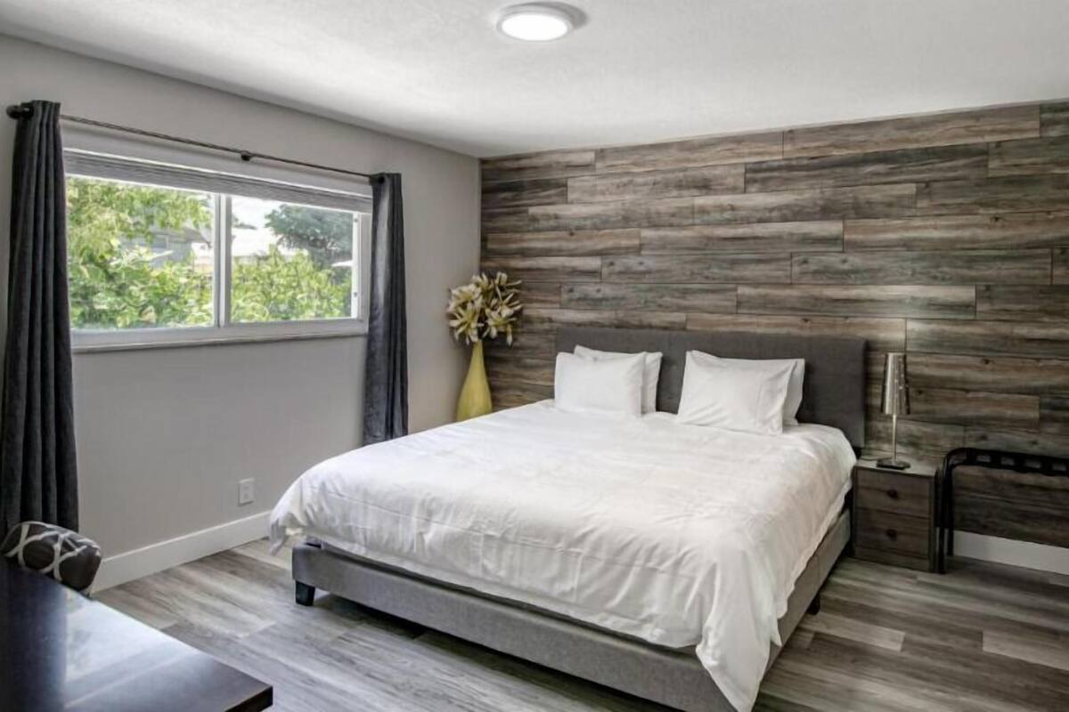 A modern bedroom with a large bed, a wooden accent wall, a window with curtains, and a bedside table with a lamp.