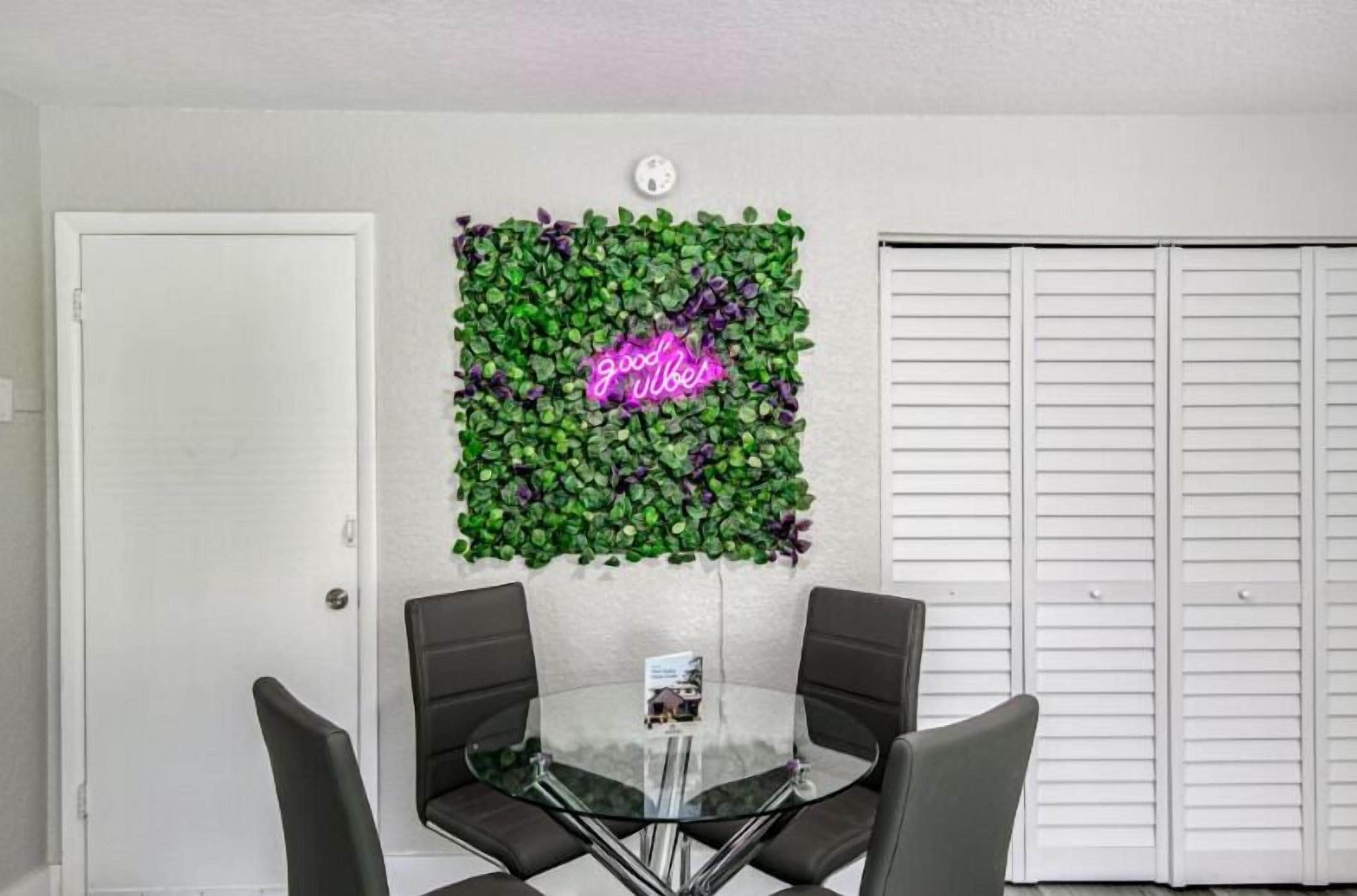 Una habitación con una mesa de vidrio y cuatro sillas, un letrero de neón sobre una pared cubierta de hojas verdes, y una puerta blanca.