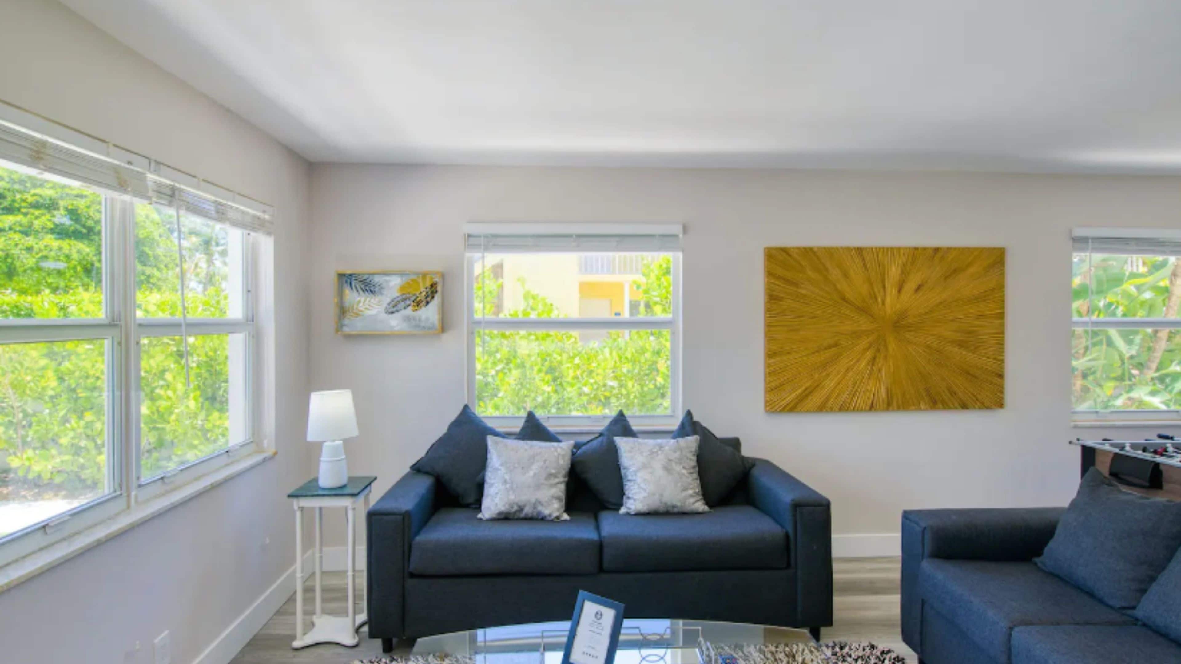 A modern living room with a dark blue sofa, a glass coffee table, and a large window providing a view of greenery outside.