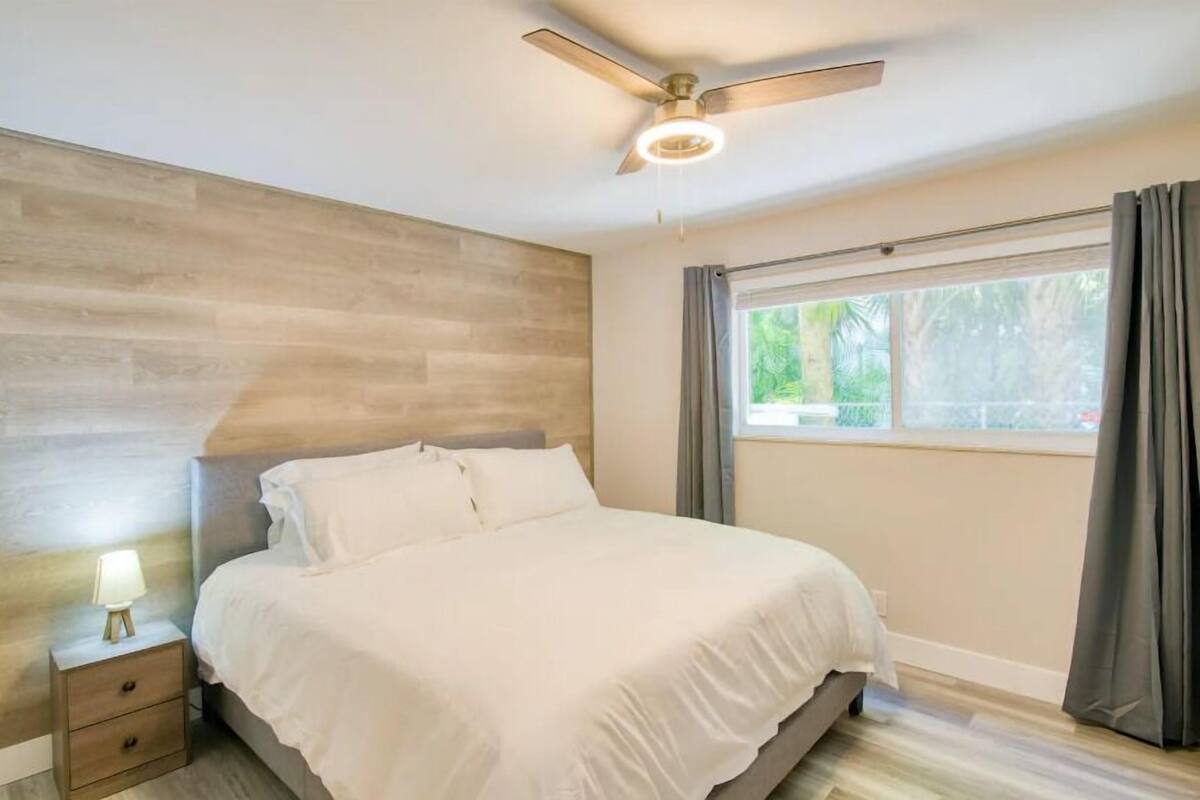 A bedroom with a bed, a nightstand with a lamp, a ceiling fan, and a window with curtains.