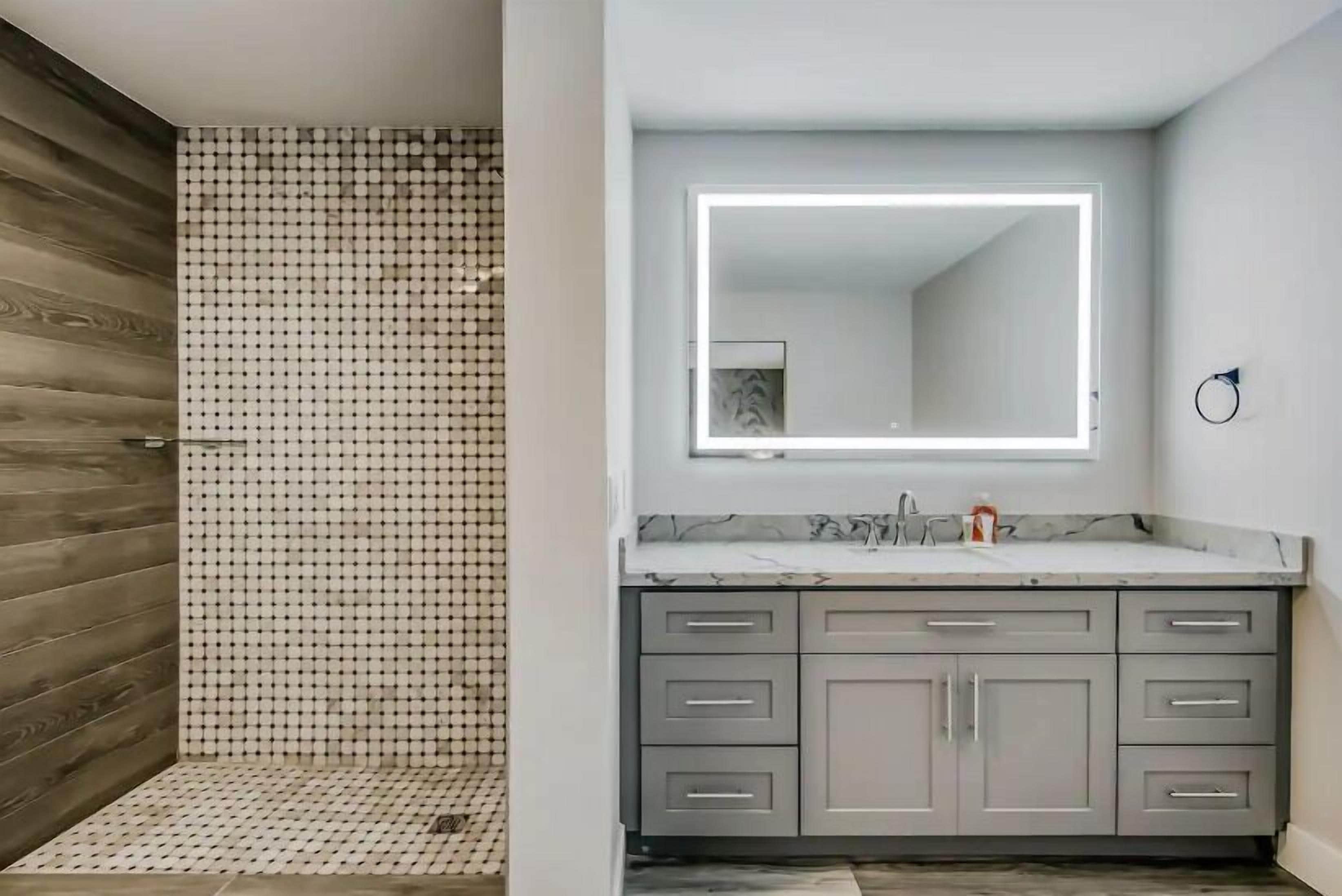 A modern bathroom with a shower area featuring a patterned tile wall and floor, a large mirror with integrated lighting, and a grey vanity with multiple drawers.