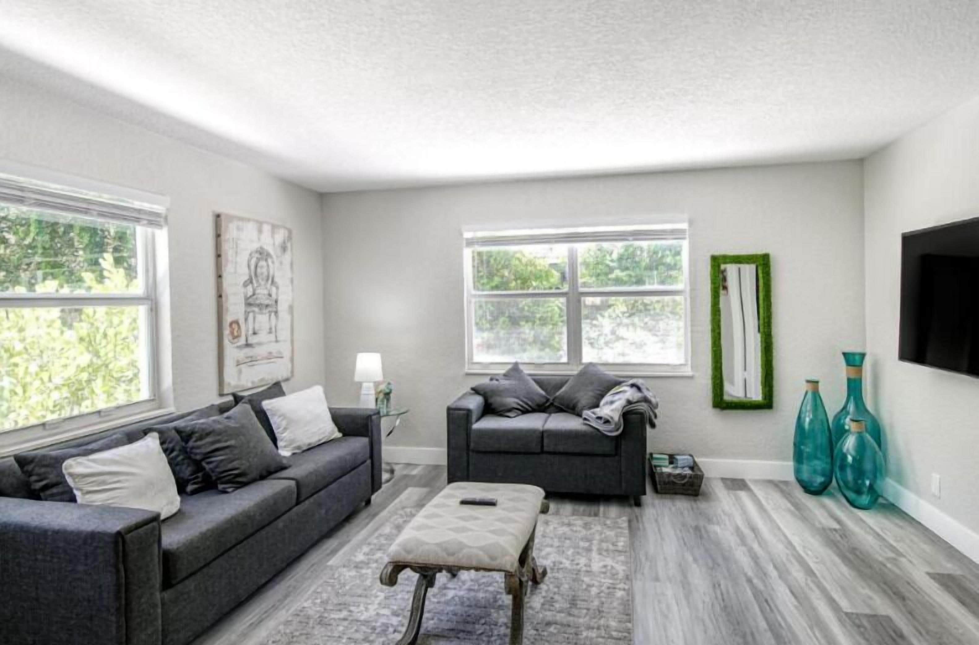 A living room with a grey sofa, a small ottoman, and a coffee table. There are two windows with blinds, a green-framed mirror, and teal vases on the floor.