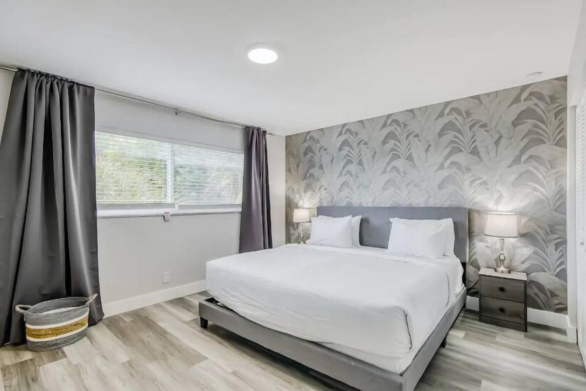 A modern bedroom with a large bed, a nightstand with a lamp, and a window with curtains.