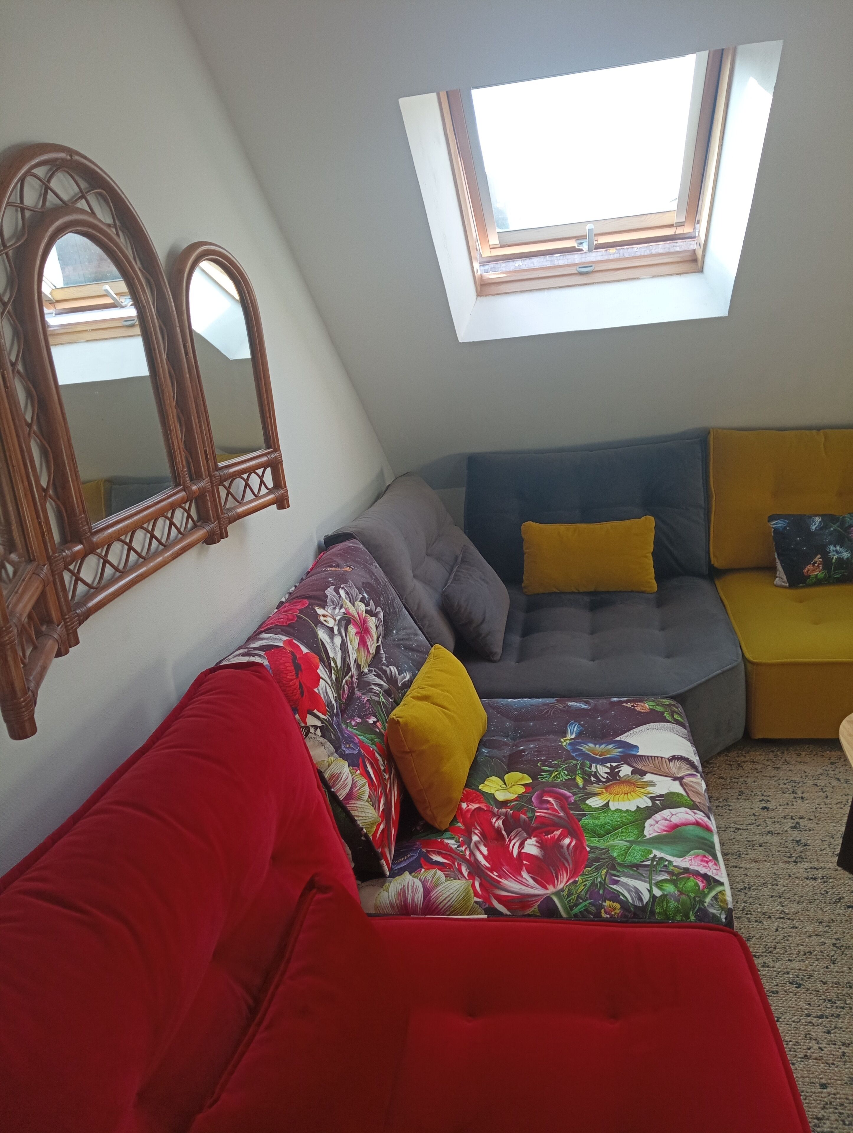 A cozy room with a sloped ceiling, a large window, a red sofa, a grey couch, and a yellow ottoman.