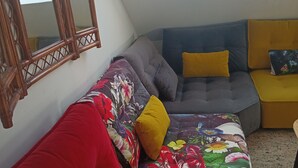 Une pièce chaleureuse avec un plafond en pente, une grande fenêtre, un canapé rouge, un divan gris et un pouf jaune.