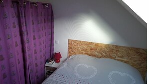 Une chambre avec un plafond en pente, une tête de lit en bois, un lit orné de broderies en forme de cœur, une table de chevet avec une lampe rouge et des rideaux violets à motifs.