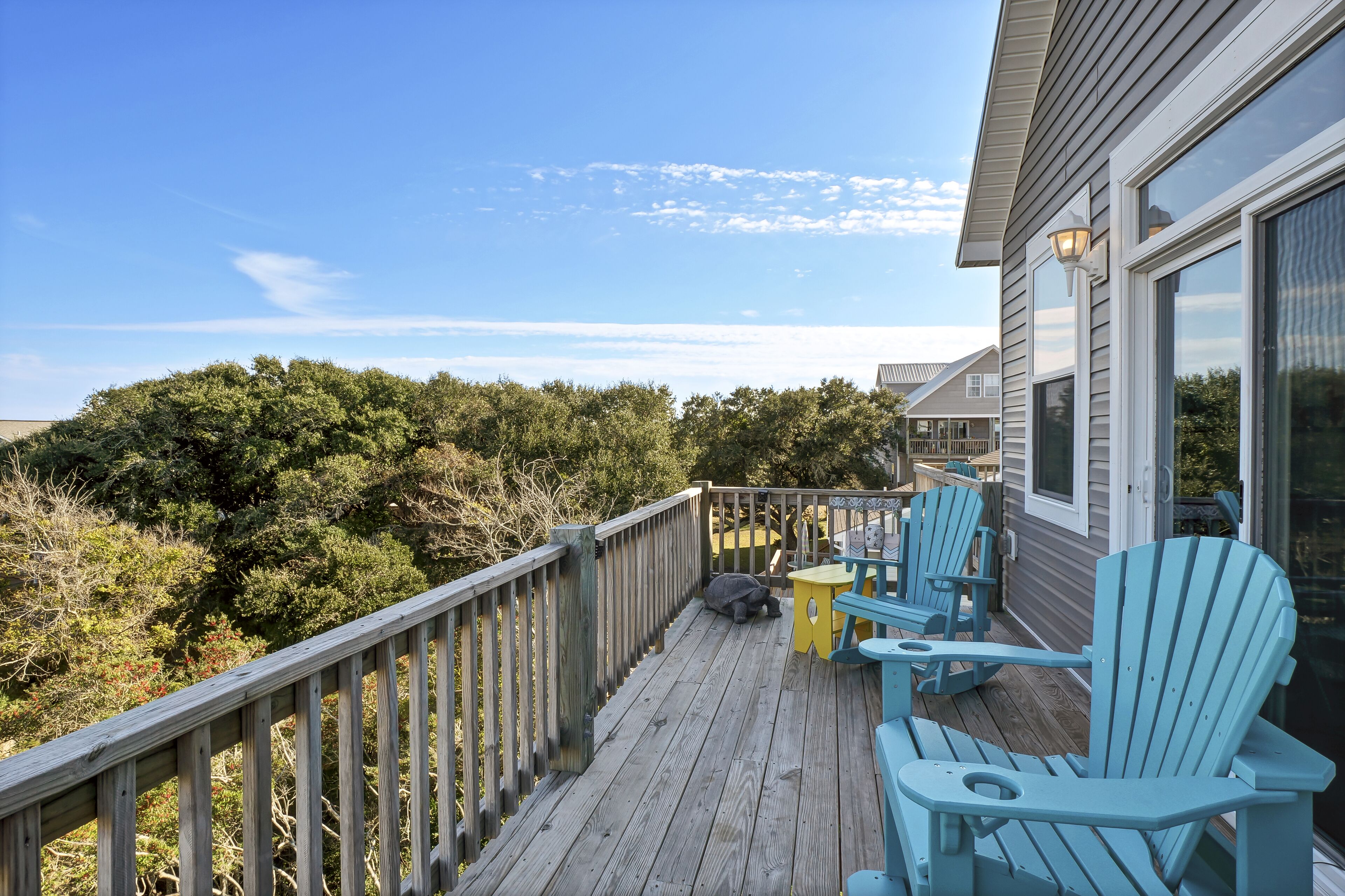 A wooden deck with blue Adirondack chairs and a yellow table, overlooking a forested area.