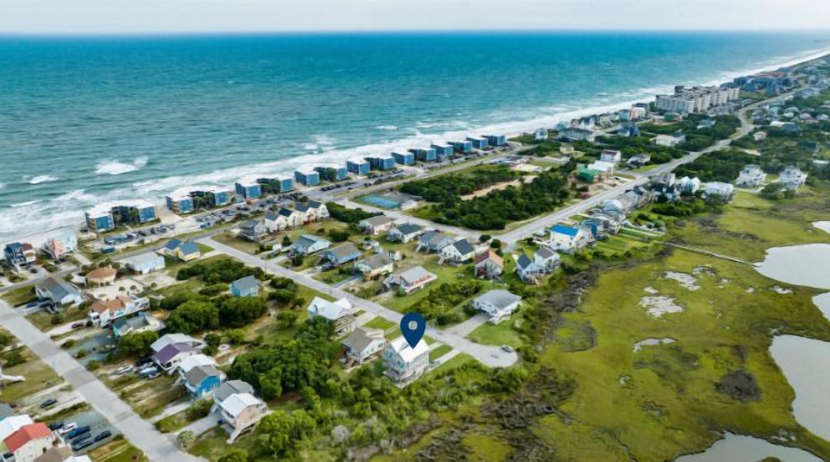 An aerial view of a coastal residential area with houses and buildings along the beachfront, a clear view of the ocean, and a marked location pin.