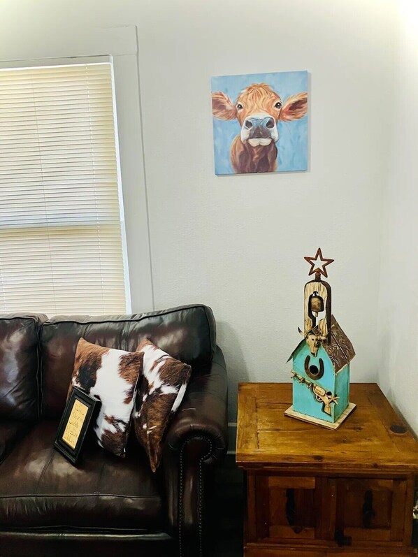A living room with a brown leather couch, a framed picture of a cow, and a decorative wooden horsehouse with a cow skull on top.