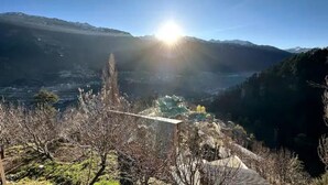 A mountainous landscape with a clear sky and the sun shining. There are trees and a small building visible in the foreground.