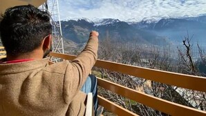 A person seated on a balcony overlooking a mountainous landscape with snow-capped peaks and a body of water.