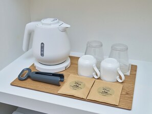 Une bouilloire électrique blanche, deux tasses blanches, deux verres, un rouleau à pâtisserie noir et deux sacs en papier marron ornés de logos, le tout disposé sur une planche en bois posée sur une table blanche.