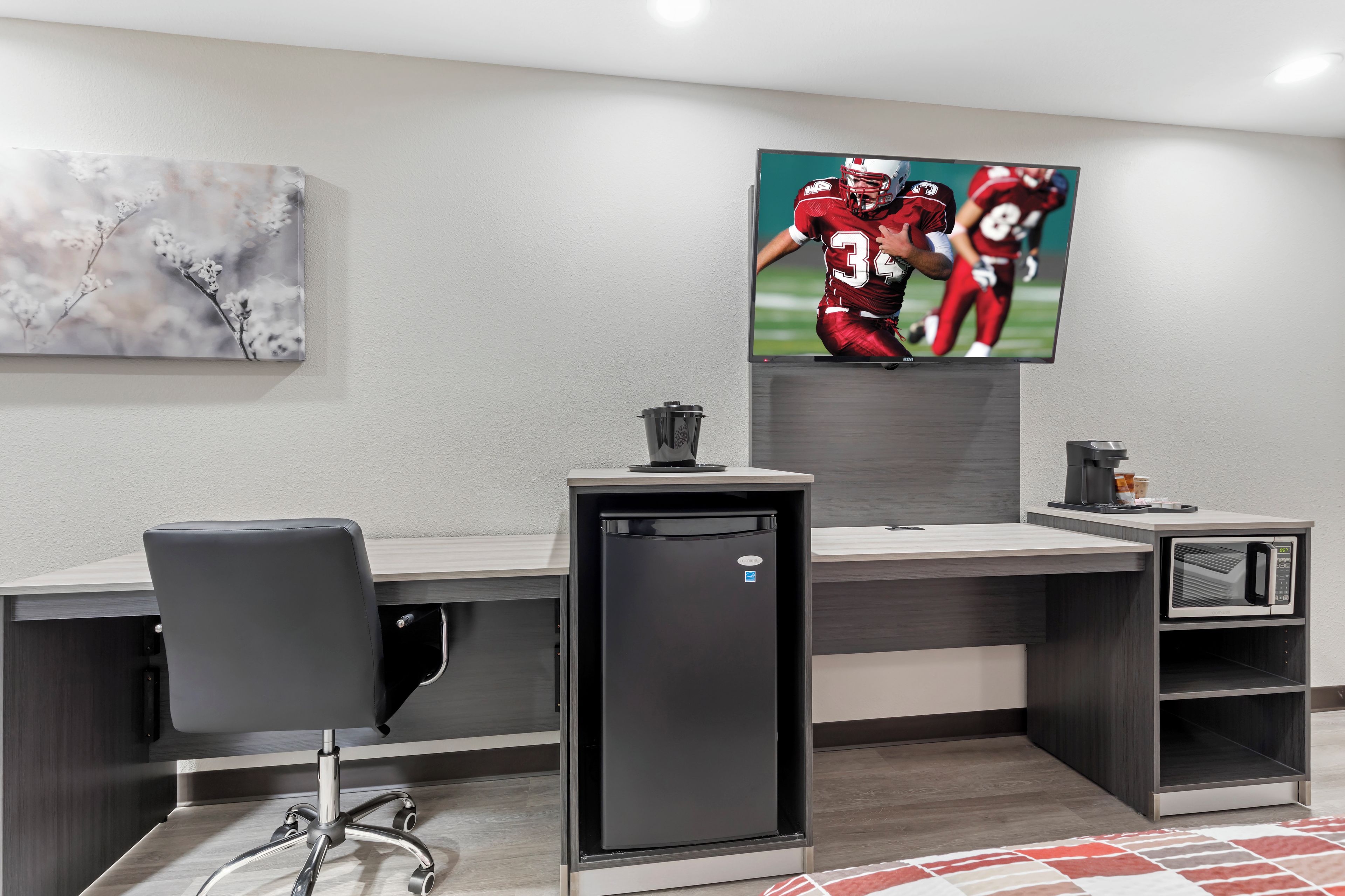 A modern office space with a desk, chair, refrigerator, and a flat-screen TV mounted on the wall.