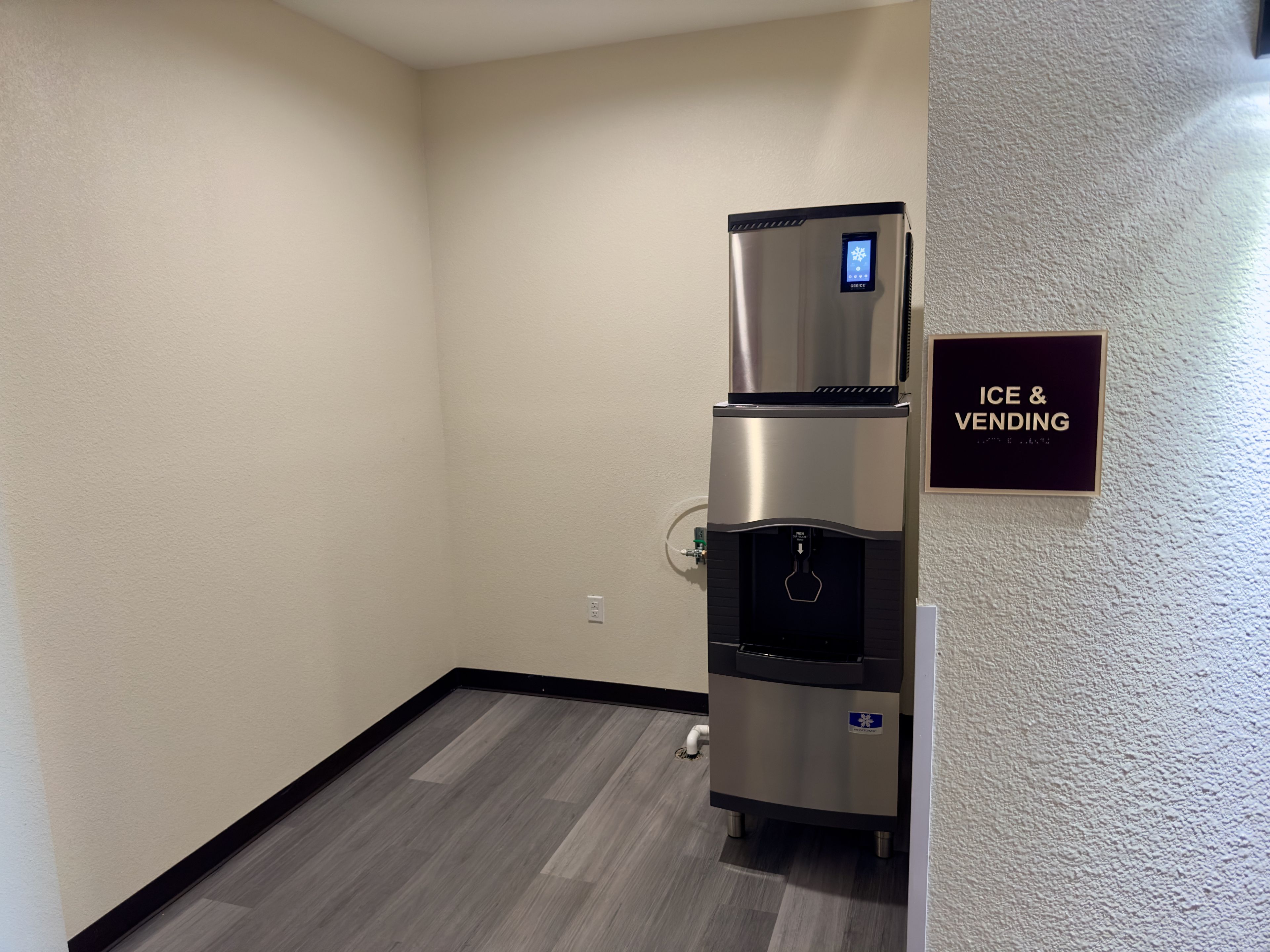 A kitchenette with a stainless steel refrigerator and a water dispenser, a wall-mounted sign indicating 'ICE & VENDING', and a wooden floor.