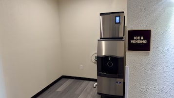 A kitchenette with a stainless steel refrigerator and a water dispenser, a wall-mounted sign indicating 'ICE & VENDING', and a wooden floor.